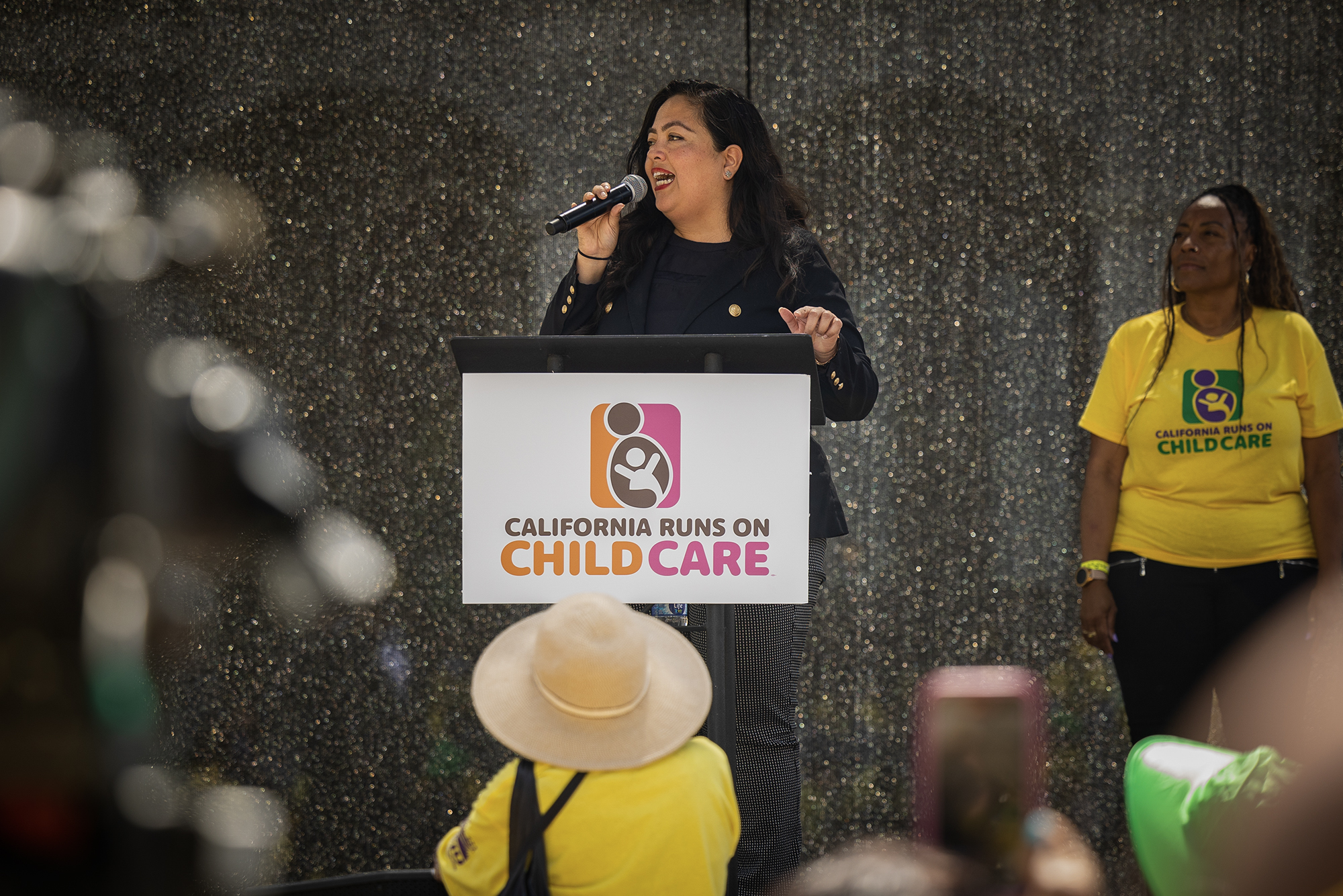 A woman wearing black clothing and holding a microphone in front of a podium that says "California Runs on Child Care."