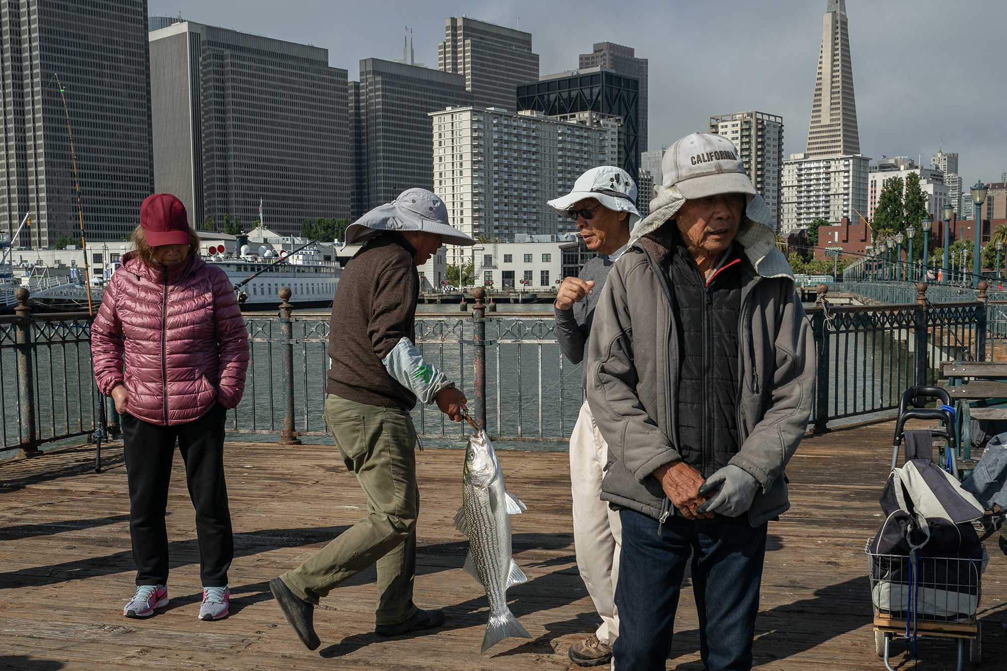 Three men and a woman wearing fishing hats and dark clothing stand on a pier with one man walking away while holding a fish.