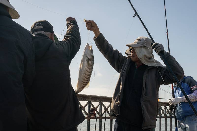 Two men hold a fishing line with a fish attached at the end of the hook with other men around with fishing poles on a pier.