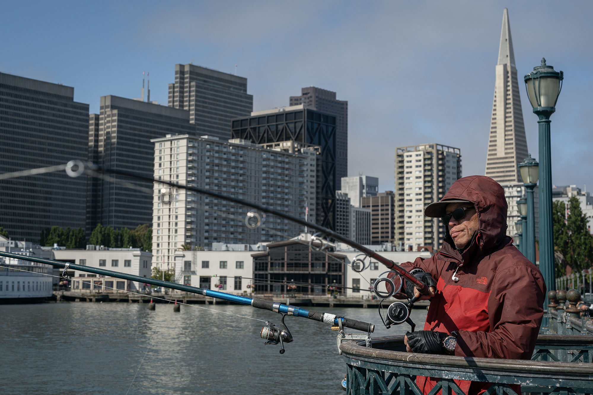 A man wearing a fishing hat and red jacket holds a fishing pole over the side of a piece with a city in the background.