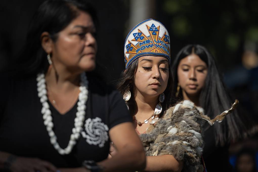 A closeup of three indigenous women standing by each other, with the woman in the middle wearing an elaborately decorated crown.