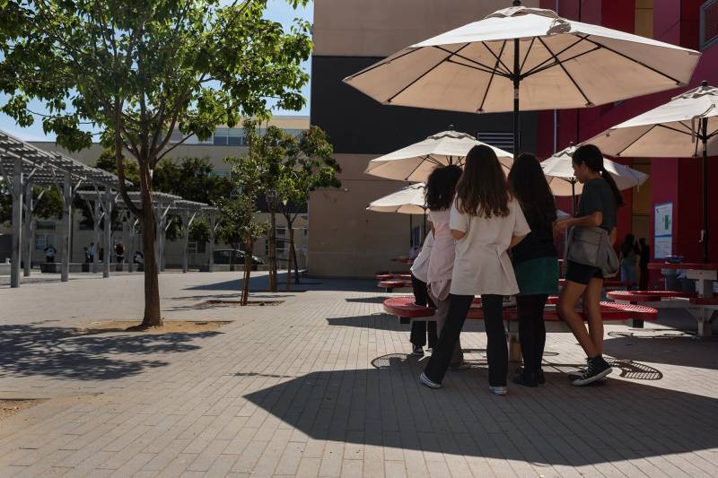 Children sit in a schoolyard at a table under a white umbrella.