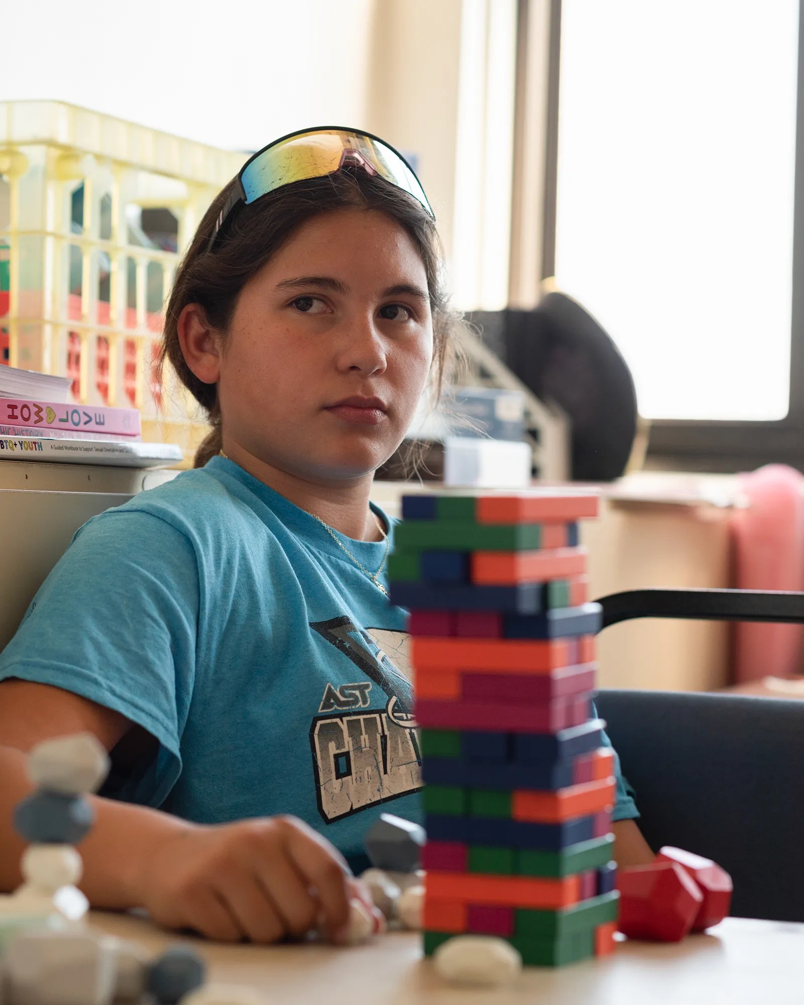 A young girl with sunglasses on her forehead sits in a classroom behind a tower of small colored blocks.