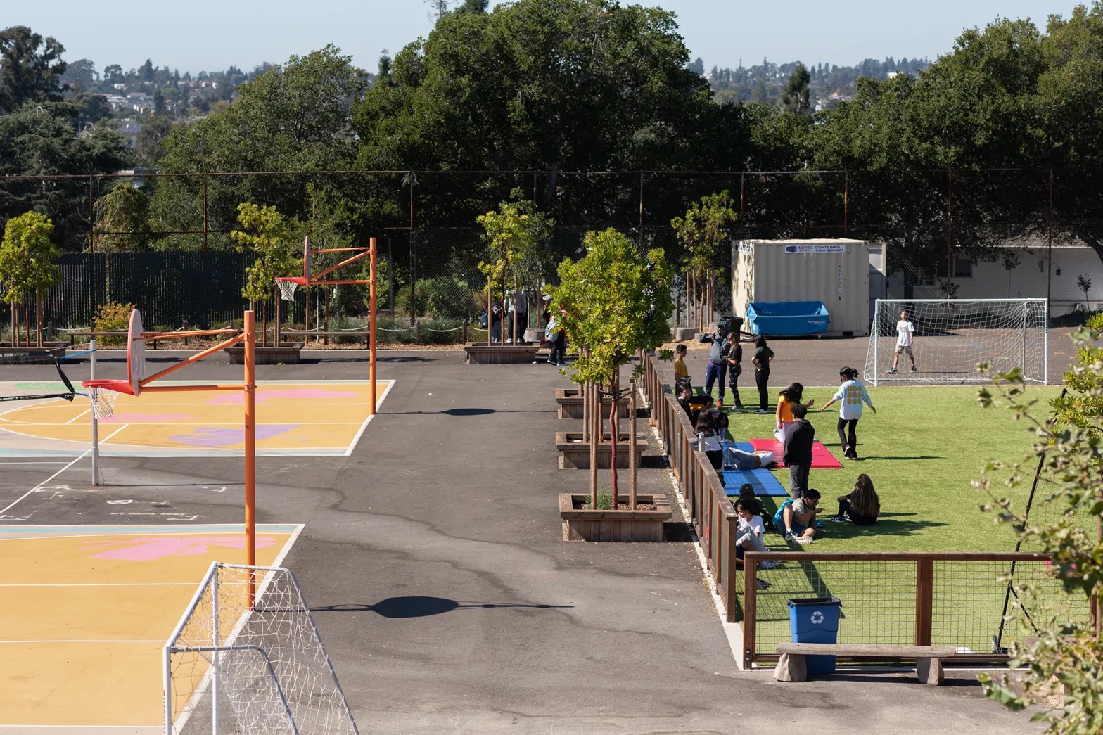 Students sit in the shade on a soccer field in a schoolyard, next to basketball courts.