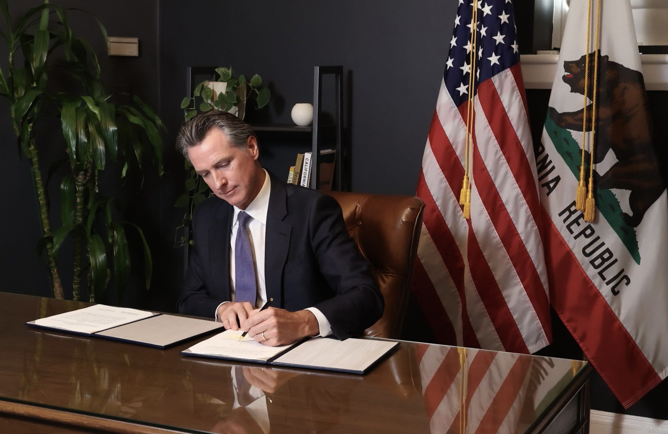 A white man wearing a suit sits at a desk in an office writing on a piece of paper with an American flag behind him.