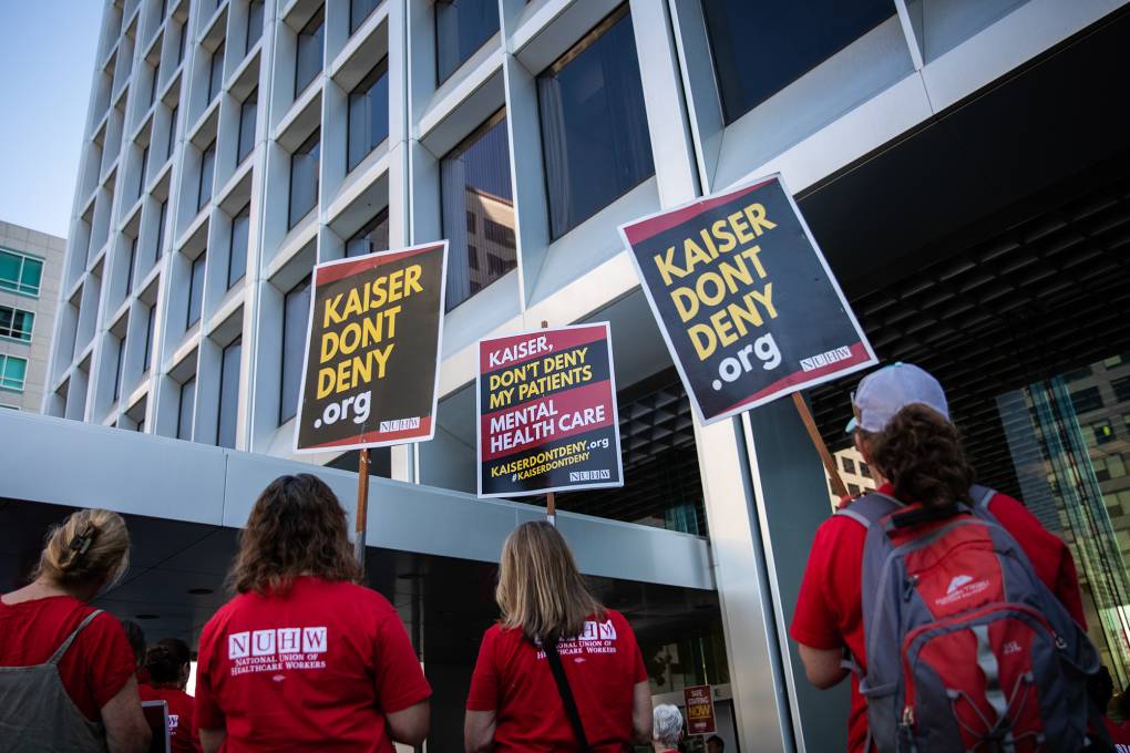 Several women wearing red shirts and holding signs that read "Kaiser Don't Deny.org" stand outside a building.
