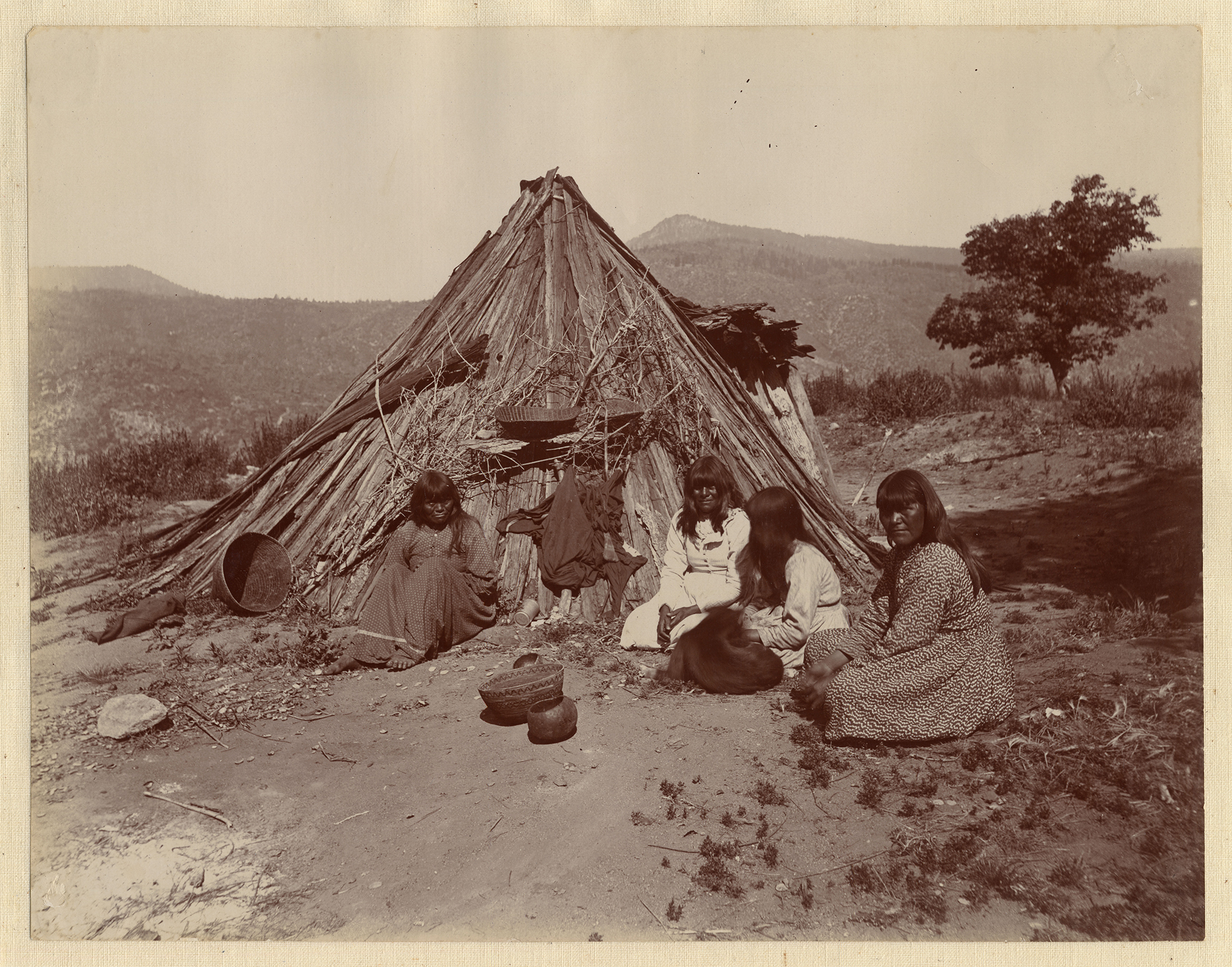 A vintage image of Native American people sitting near a bark hut.