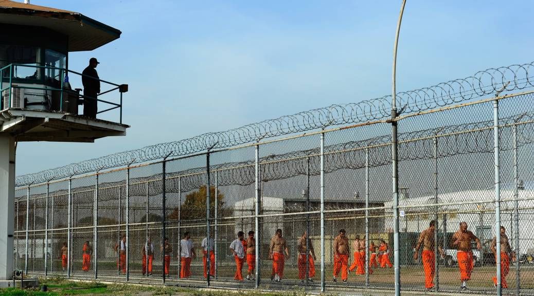 Imprisoned people wearing orange outfits hang out in an outdoor area of the prison, while a guard watches from a tower overhead.