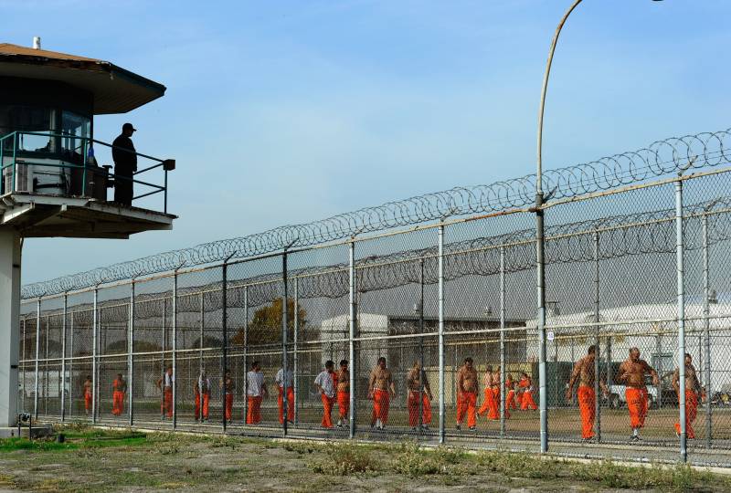 Imprisoned people wearing orange outfits hang out in an outdoor area of the prison, while a guard watches from a tower overhead.