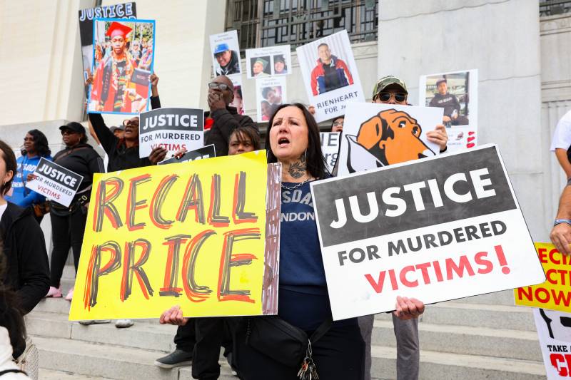 A woman at a rally in front of a courthouse holds two signs that say 'Recall Price' and 'Justice for Murder Victims.' Behind her are other people holding signs, some with photos of people on them.