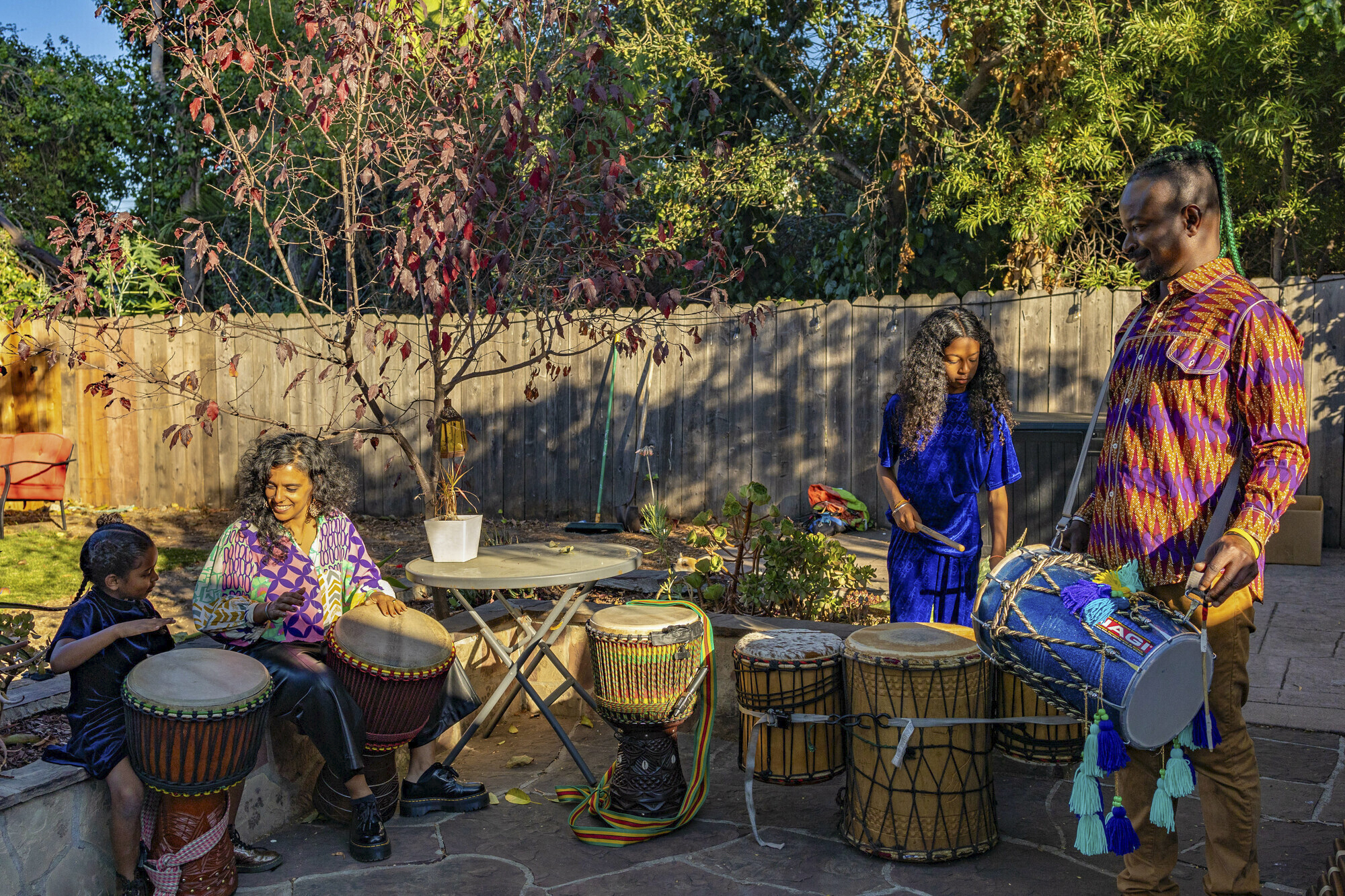 Two young mixed race girls stand and sit playing the drums next to an Indian woman and Black man.