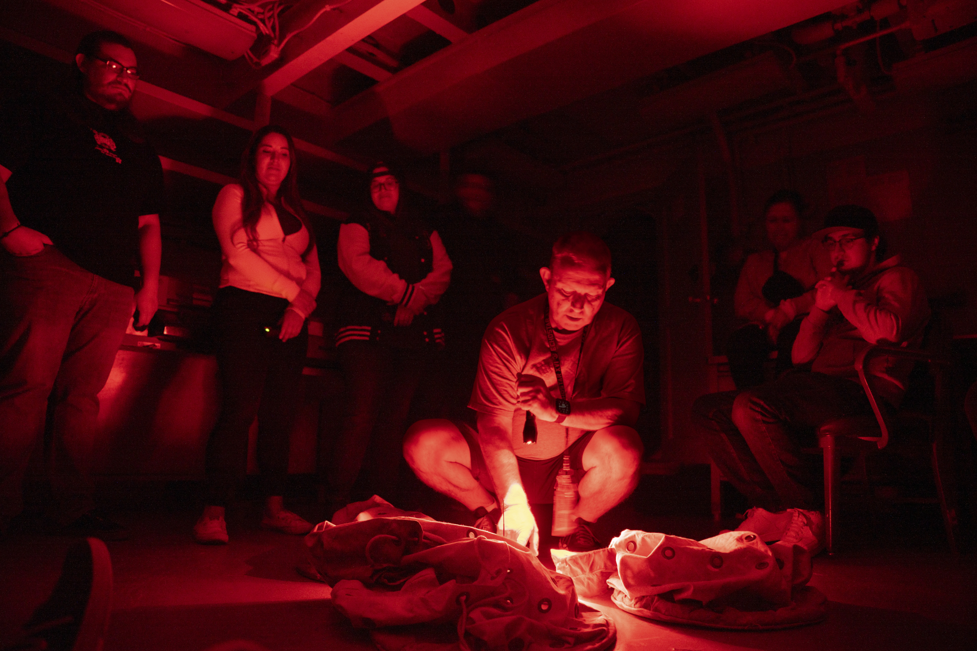 A man squats down with a measuring instrument in a red-lit room, as two other people watch.