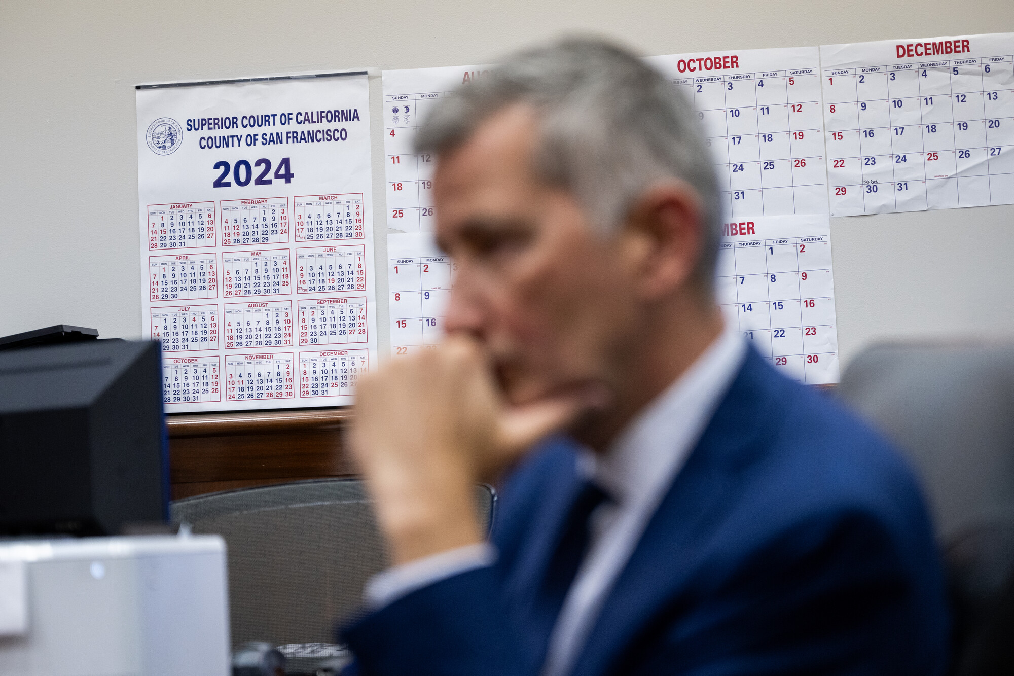 A white man wearing a blue dress suit rests his hand on his face with a calendar in the background.