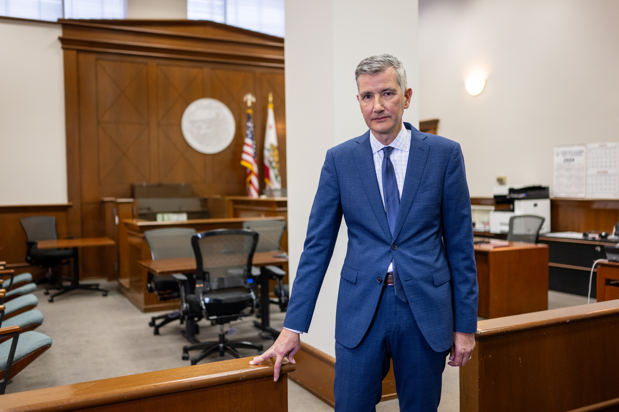 A white man wearing a blue dress suit leans against a wooden divider in a court room.