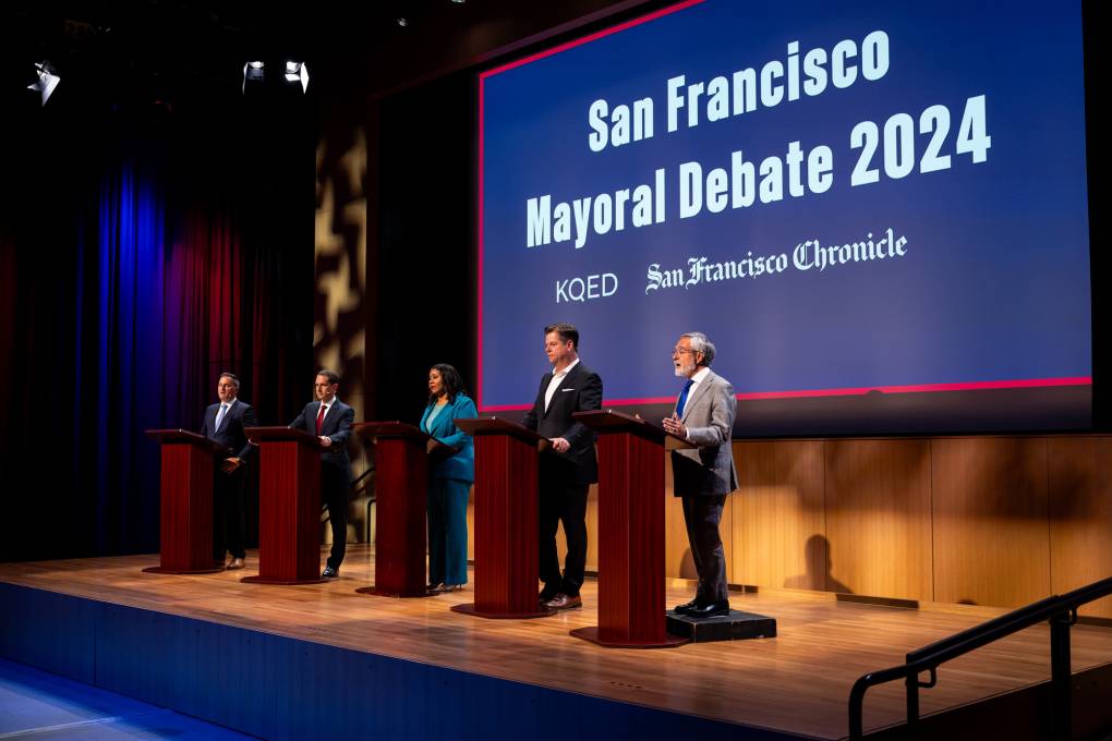 A side view of five candidates (four men and one woman), each standing behind a podium on stage, with a screen behind them that says: 'San Francisco Mayoral Debate 2024'.