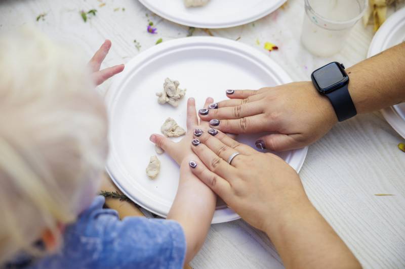An adult's hands lay on top of a little kid's hands while making something with clay.