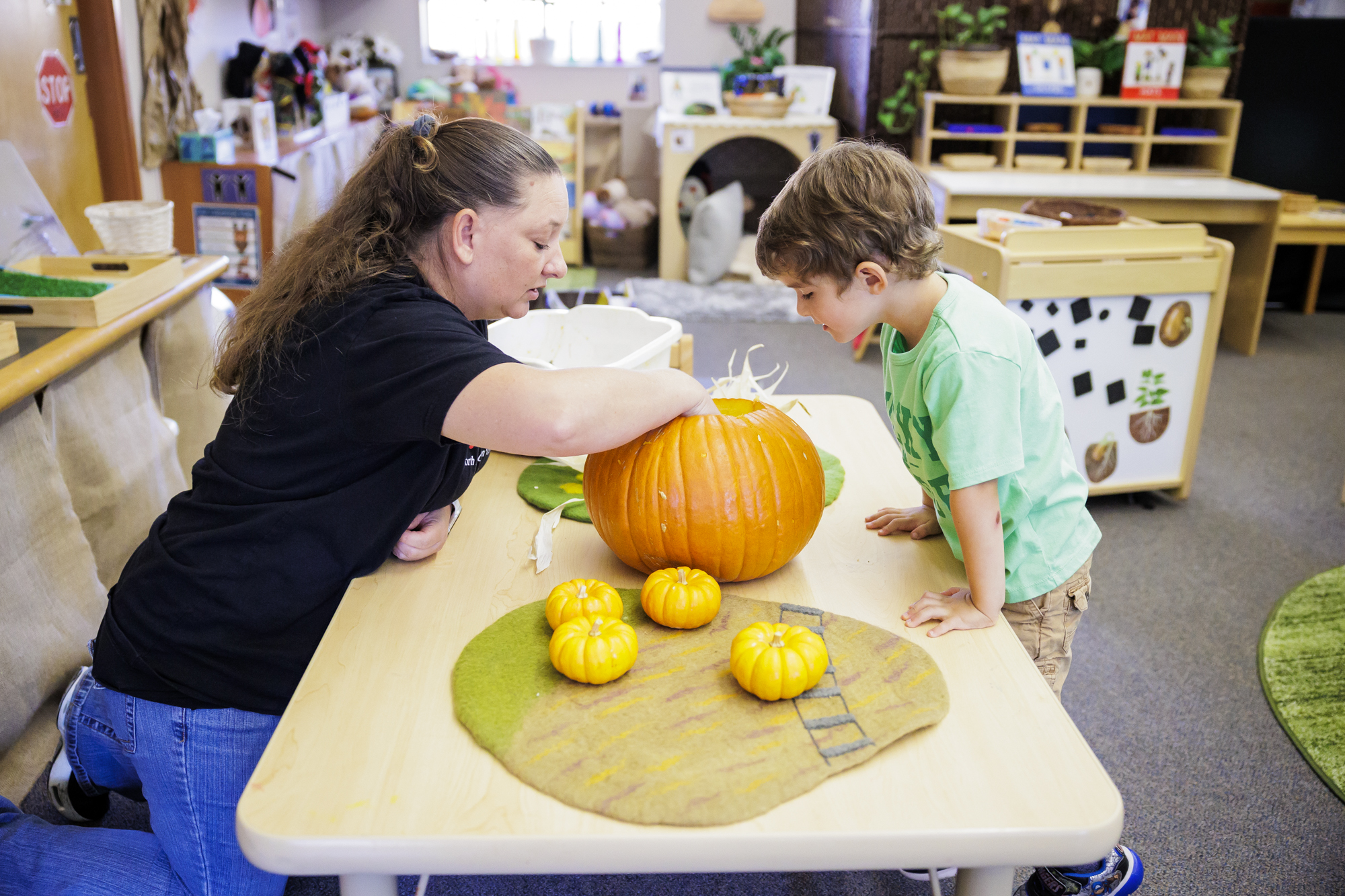 A young children standing at a table looks at a teacher digging into a pumpkin.