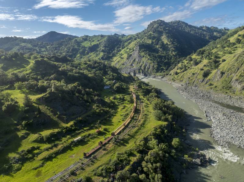 An aerial view of a landscape of mountains and forest with a railroad next to a body of water.