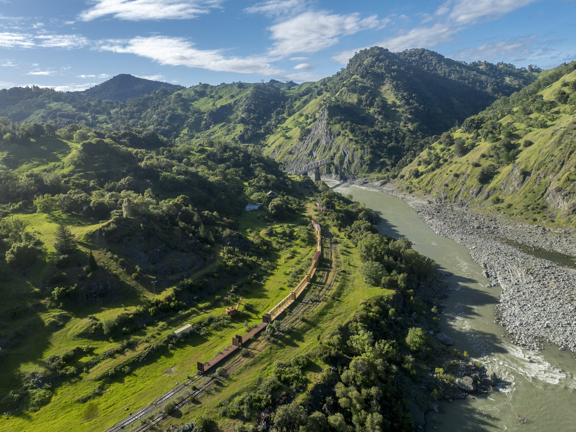 An aerial view of a landscape of mountains and forest with a railroad next to a body of water.