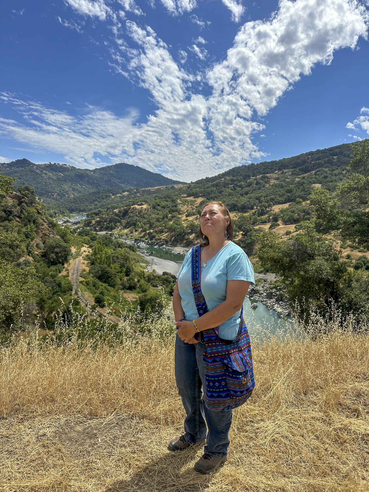 An indigenous woman wearing a turquoise shirt stands outside in nature.