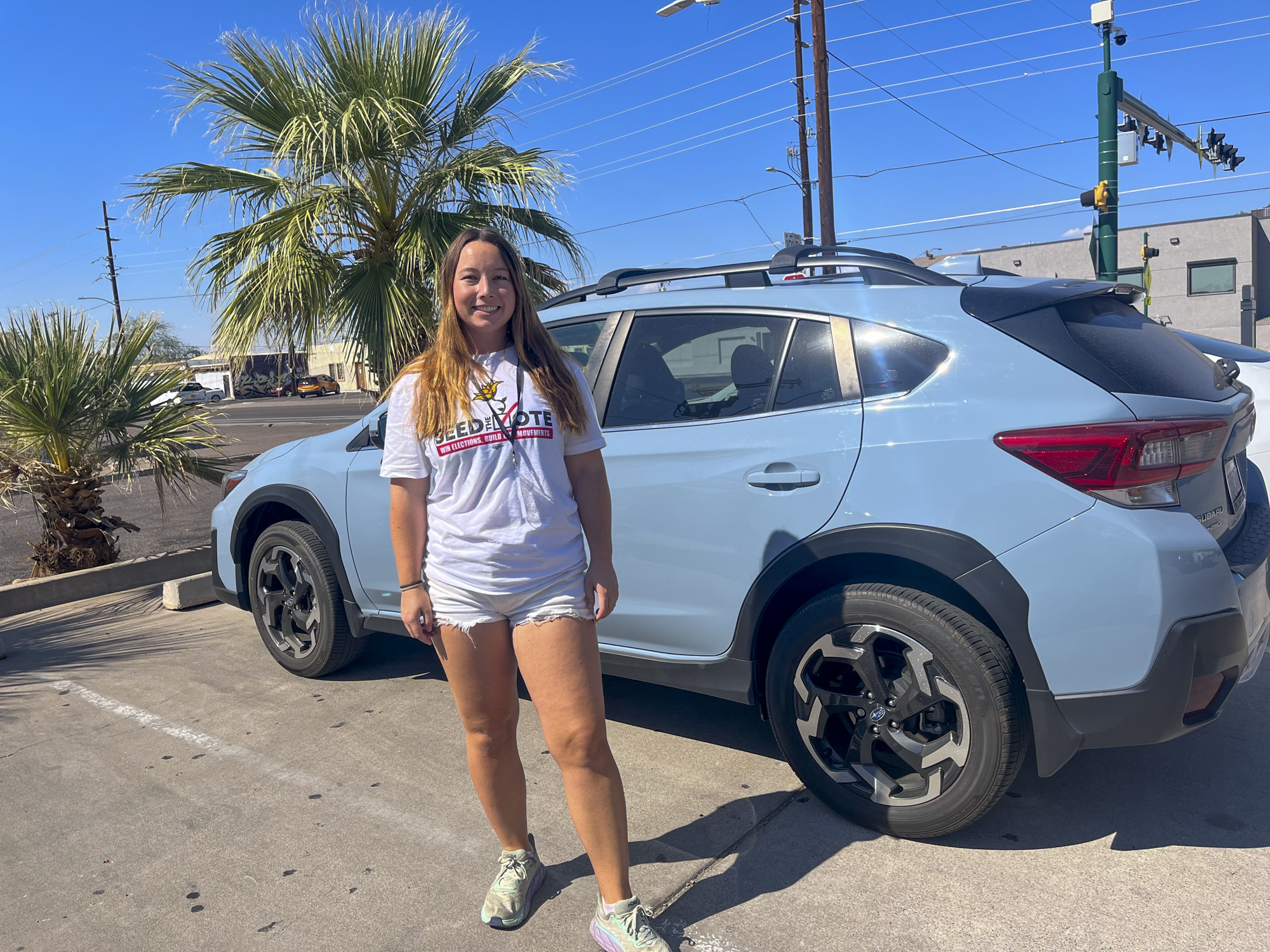 A white woman wearing a white shirt and shorts stands next to a blue car outside.