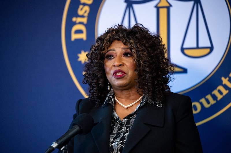 A middle-aged Black woman in a suit jacket speaks at a podium, with a 'Alameda County District Attorney's Office banner behind her.