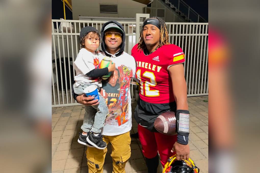 Photo of a young man holding a little boy and standing next to another young man in football uniform.