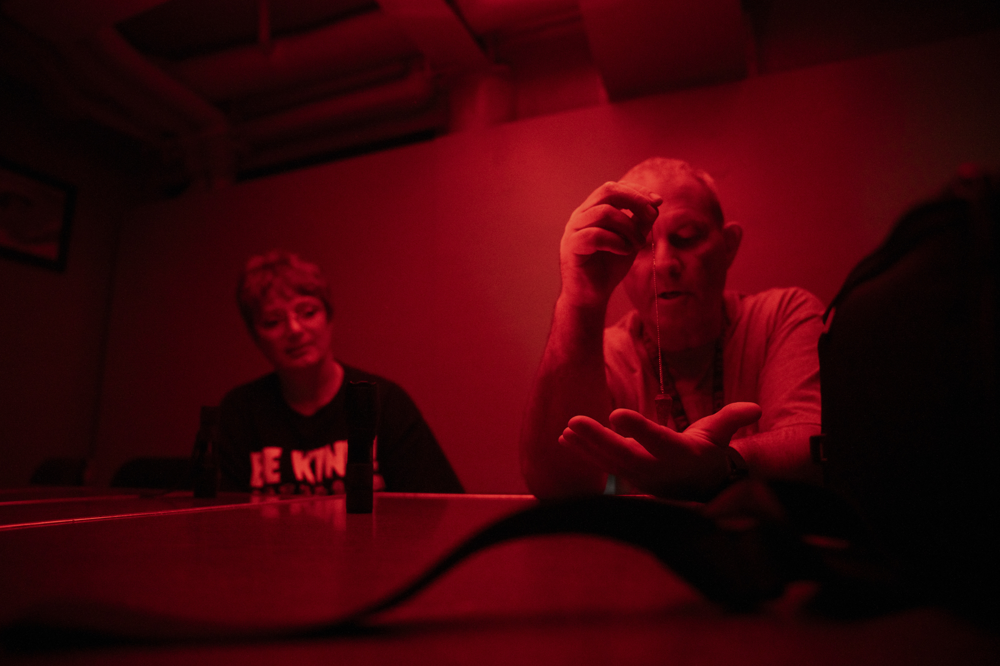A man in a red-lit room holds a small pendulum, while a woman sits next to him watching.