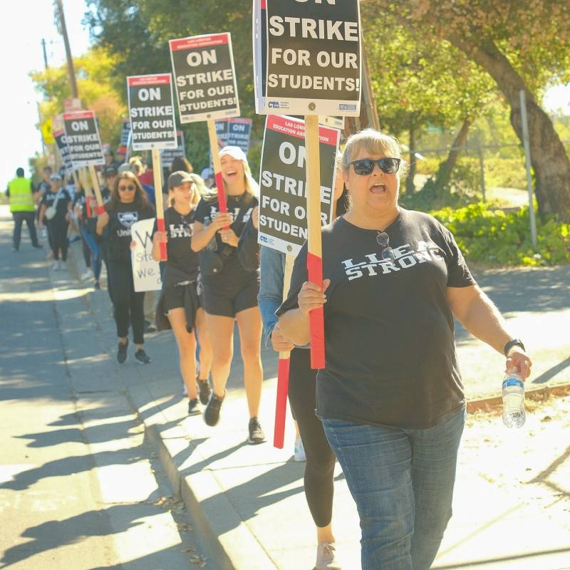 Several people wearing black T-shirts and sunglasses hold signs that read "On strike for our students" as they march down a sidewalk.