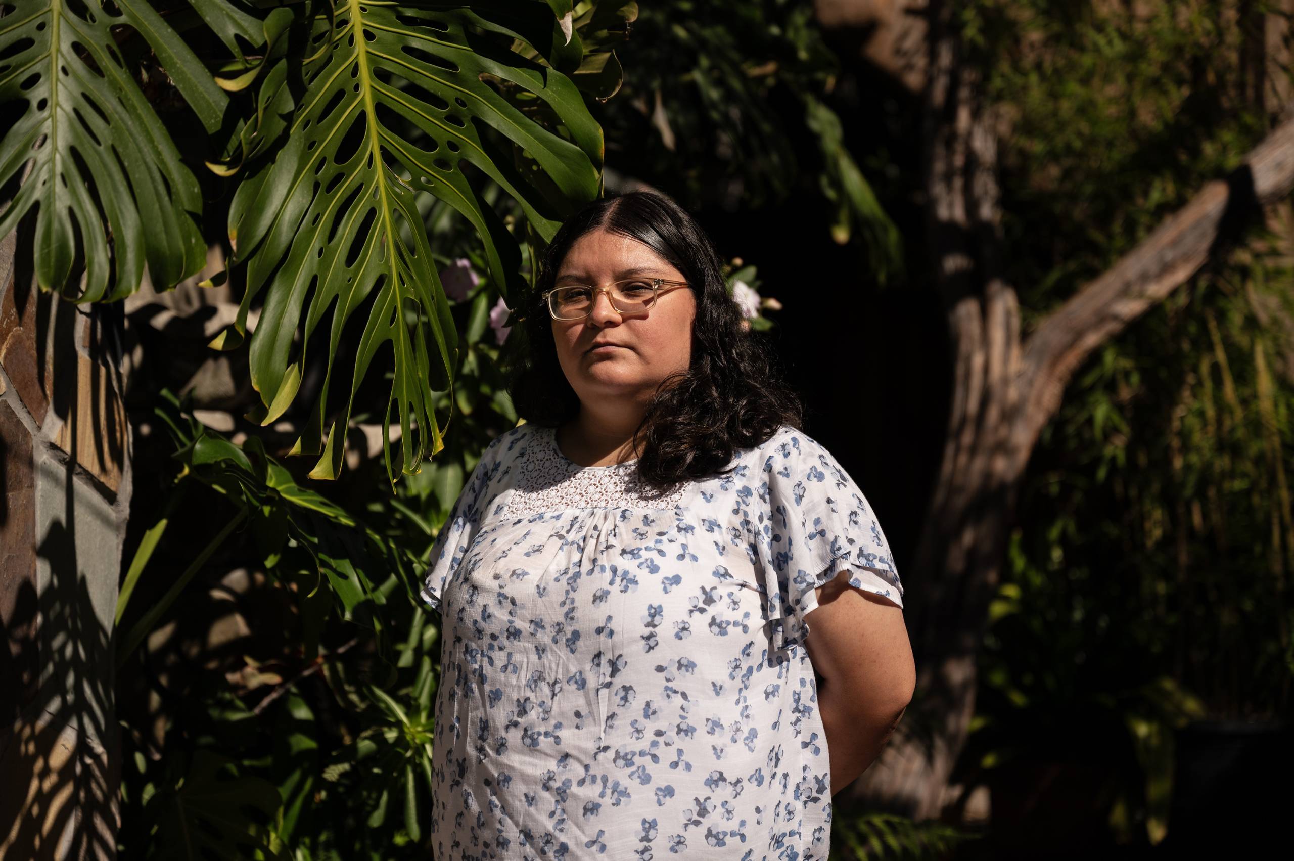 A woman with glasses and a white flowery shirt stands outside near plants.