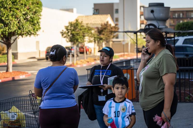 A woman in a blue shirt with a shopping cart stands near a woman holding a clipboard and a young boy. There is another woman in green holding a cellphone to her ear.