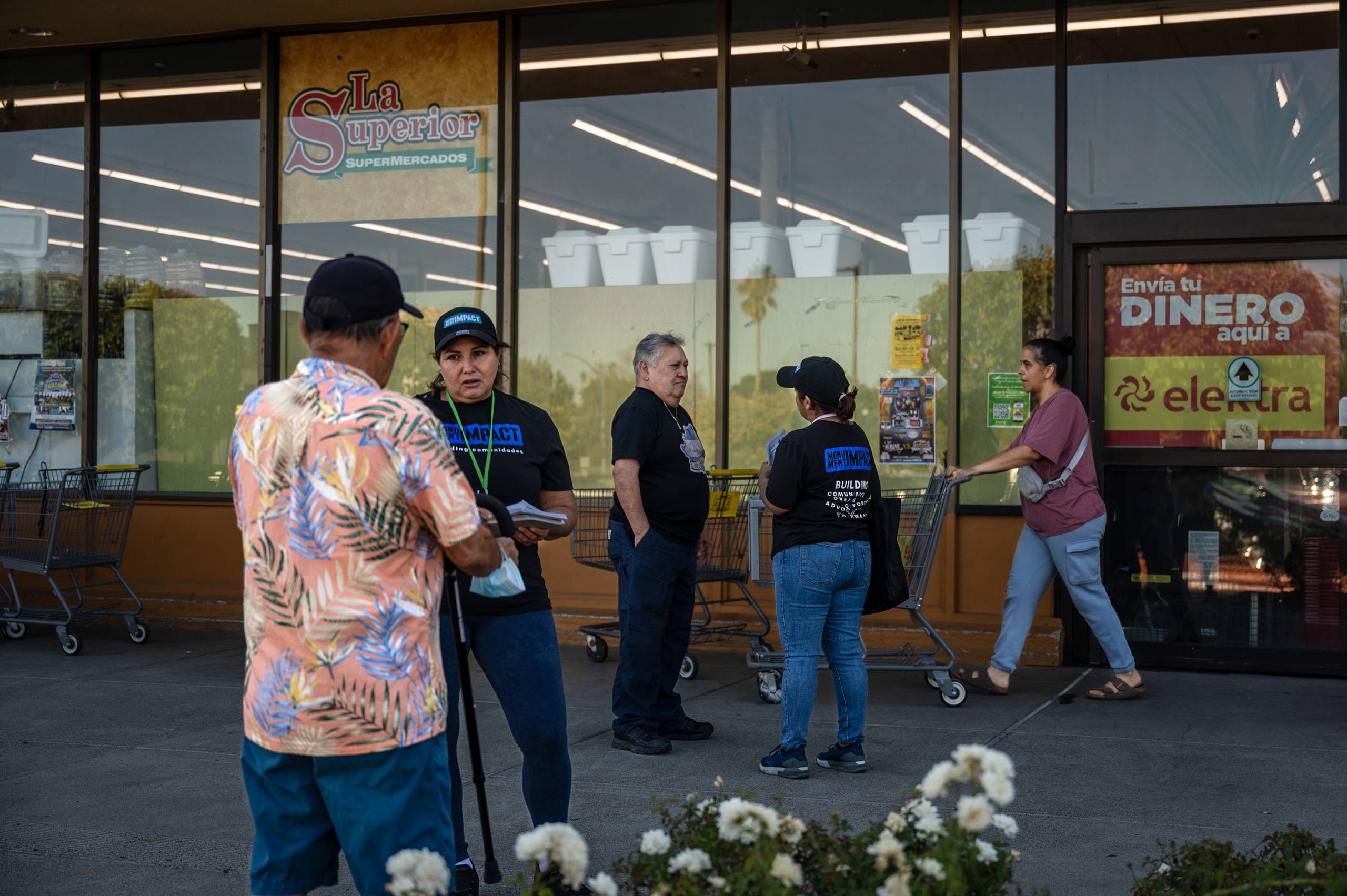 Two women wearing black t-shirts and hats talk to two men outside a building as another man walks past pushing a shopping cart.