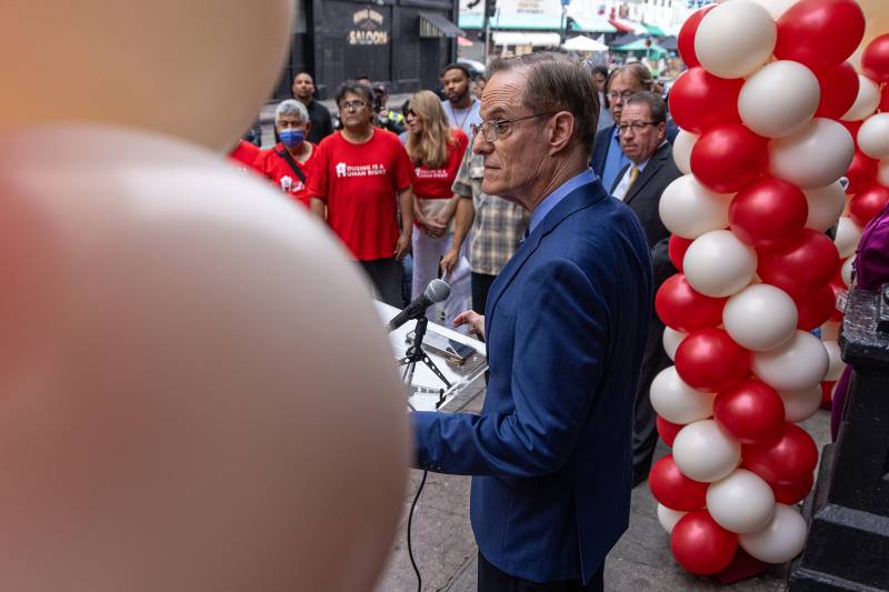 A man wearing a blue suit standing at a podium surrounded by balloons. He has a serious expression. A crowd in front of him has their eyes on him.