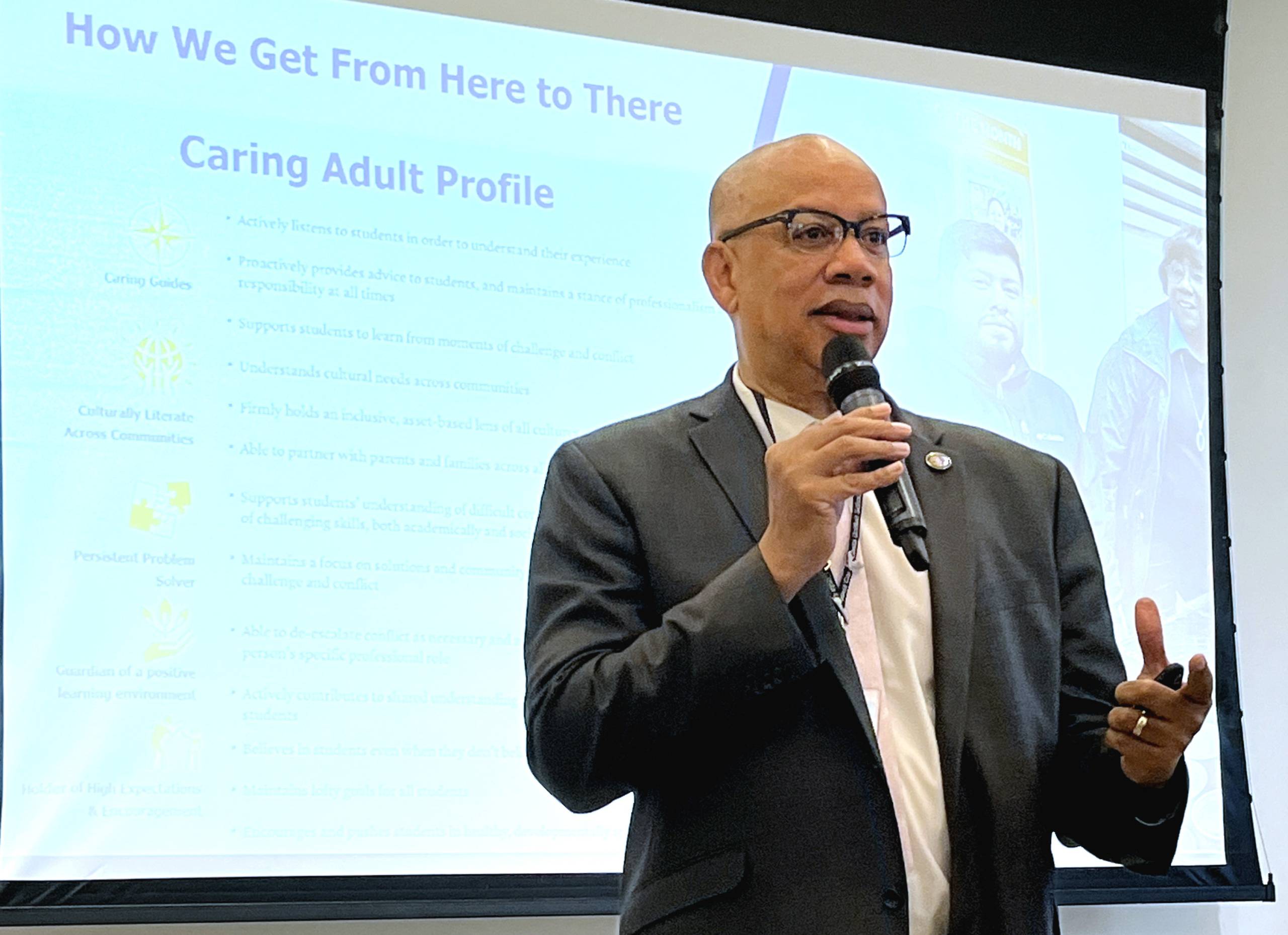 A Black man with glasses speaks into a microphone while standing in front of a large screen.