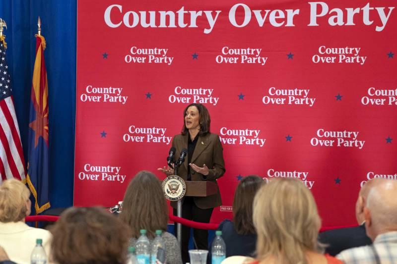 A woman wearing a brown jacket stands behind a podium with a red banner behind her that reads "Country Over Party."