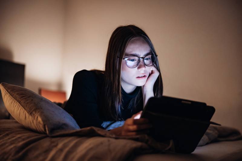 A young white girl rests on her stomach on a bed, looking at a tablet.