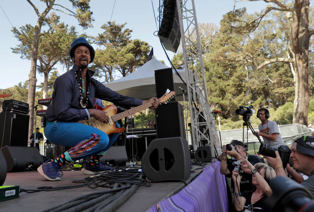 A Black man wearing blue pants and a dark shirt squats while playing a guitar on stage as people take pictures from below.