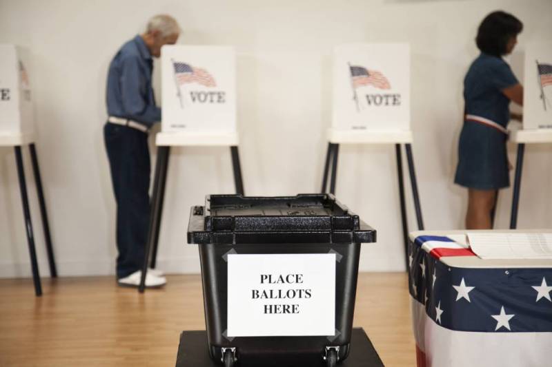 Varias personas llenan su boleta electoral dentro de cubículos de votación. Enfrente de ellos, hay una caja negra con un papel encima que dice en inglés: "Coloque sys boletas aquí".