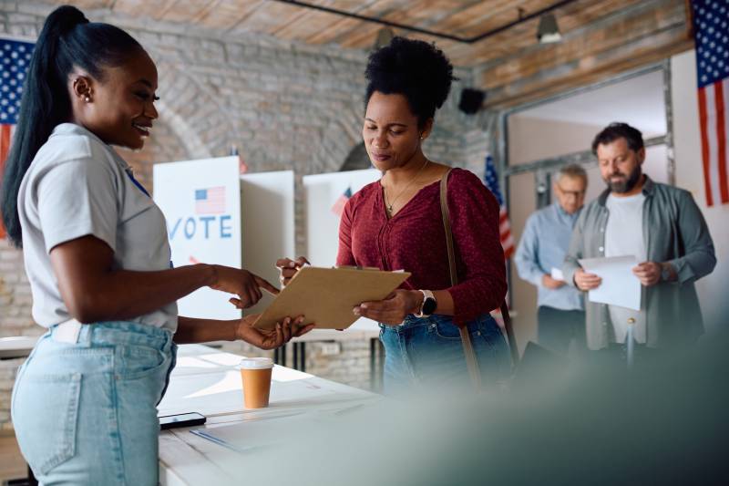 Two Black women standing and looking at a clipboard over a table and signs that say "Vote" with other people waiting in line behind the woman with the red shirt.