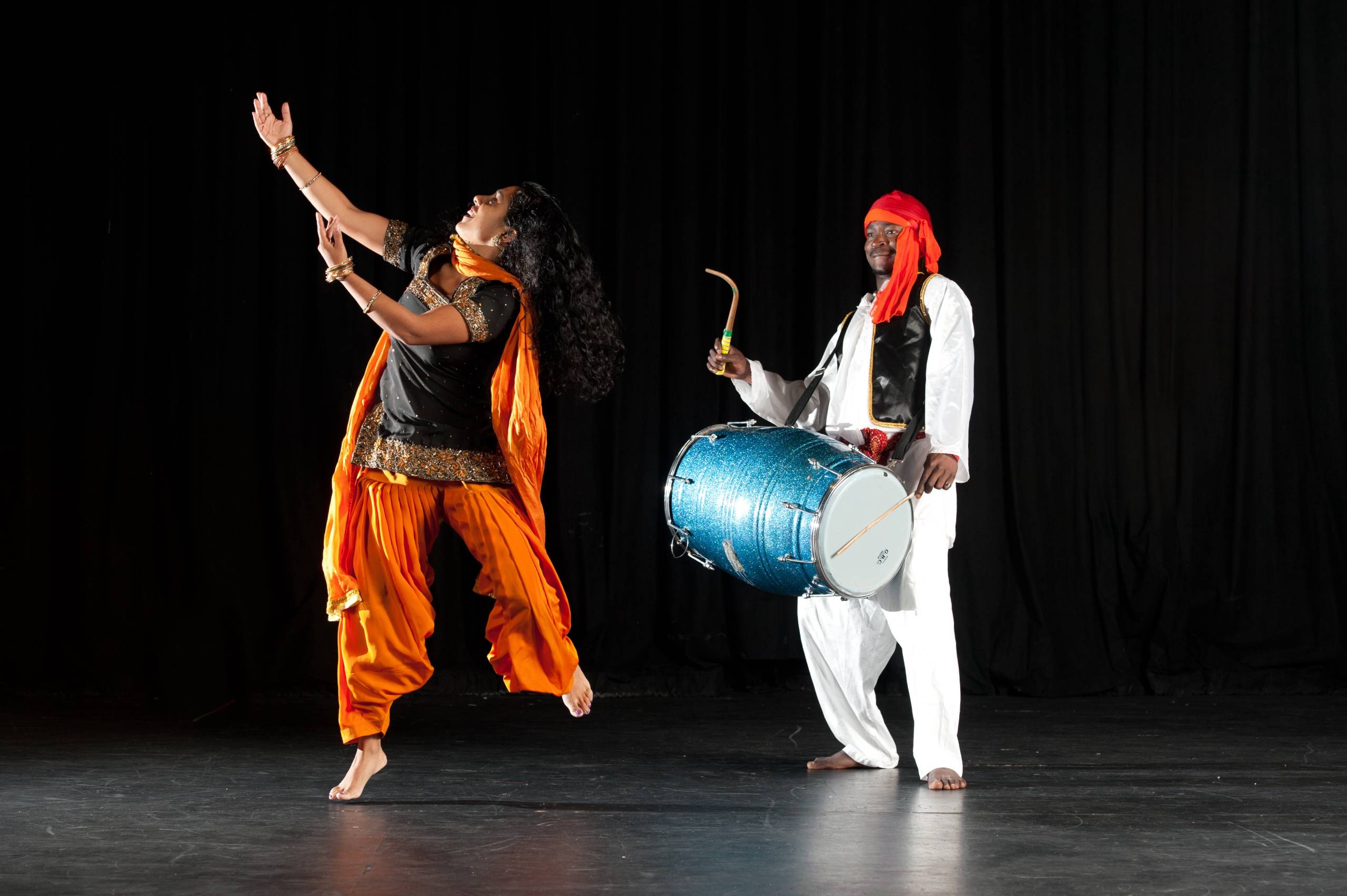 An Indian woman dressed in orange and black and a Black man dressed in white playing a drum perform on a stage.