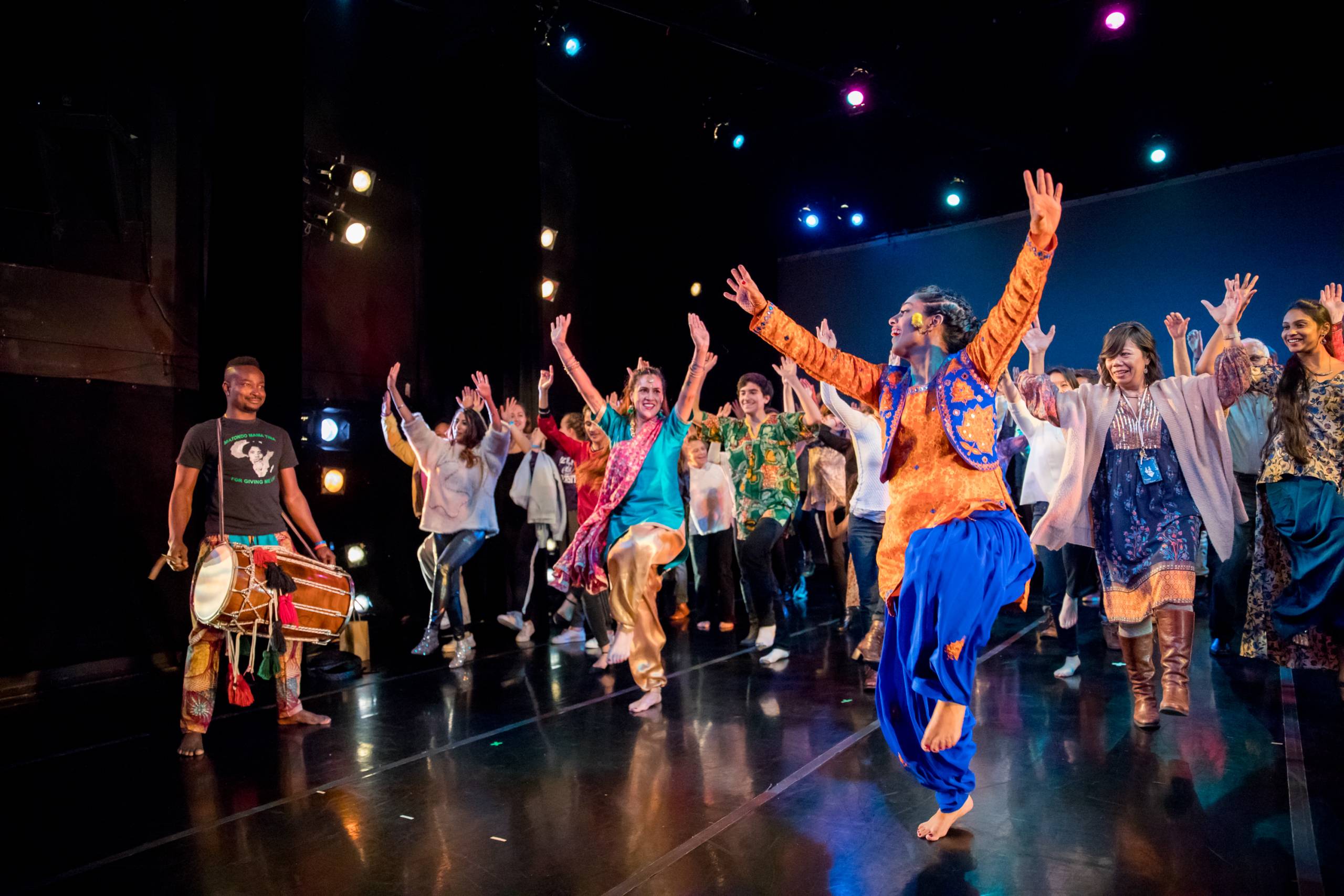 A group of mostly women wearing colorful clothing dance on a stage.