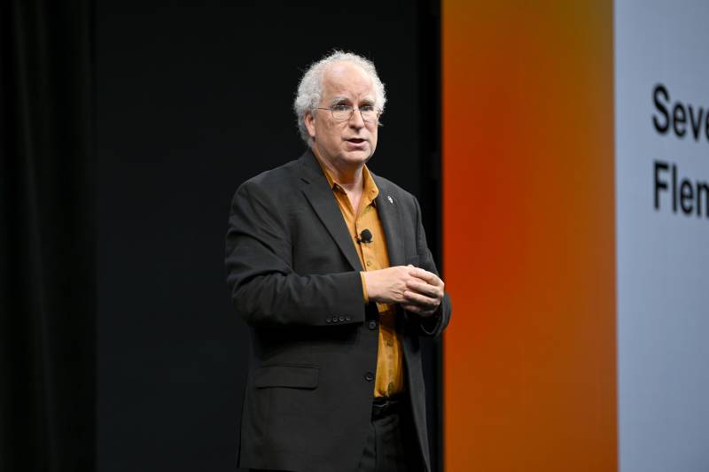 A middle-aged white man with grey hair, wearing a blazer, speaks on a stage.