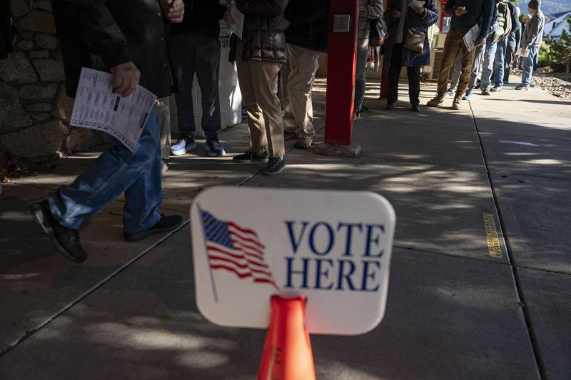 People wait in line on a street next to a sign that says 'Vote Here.'