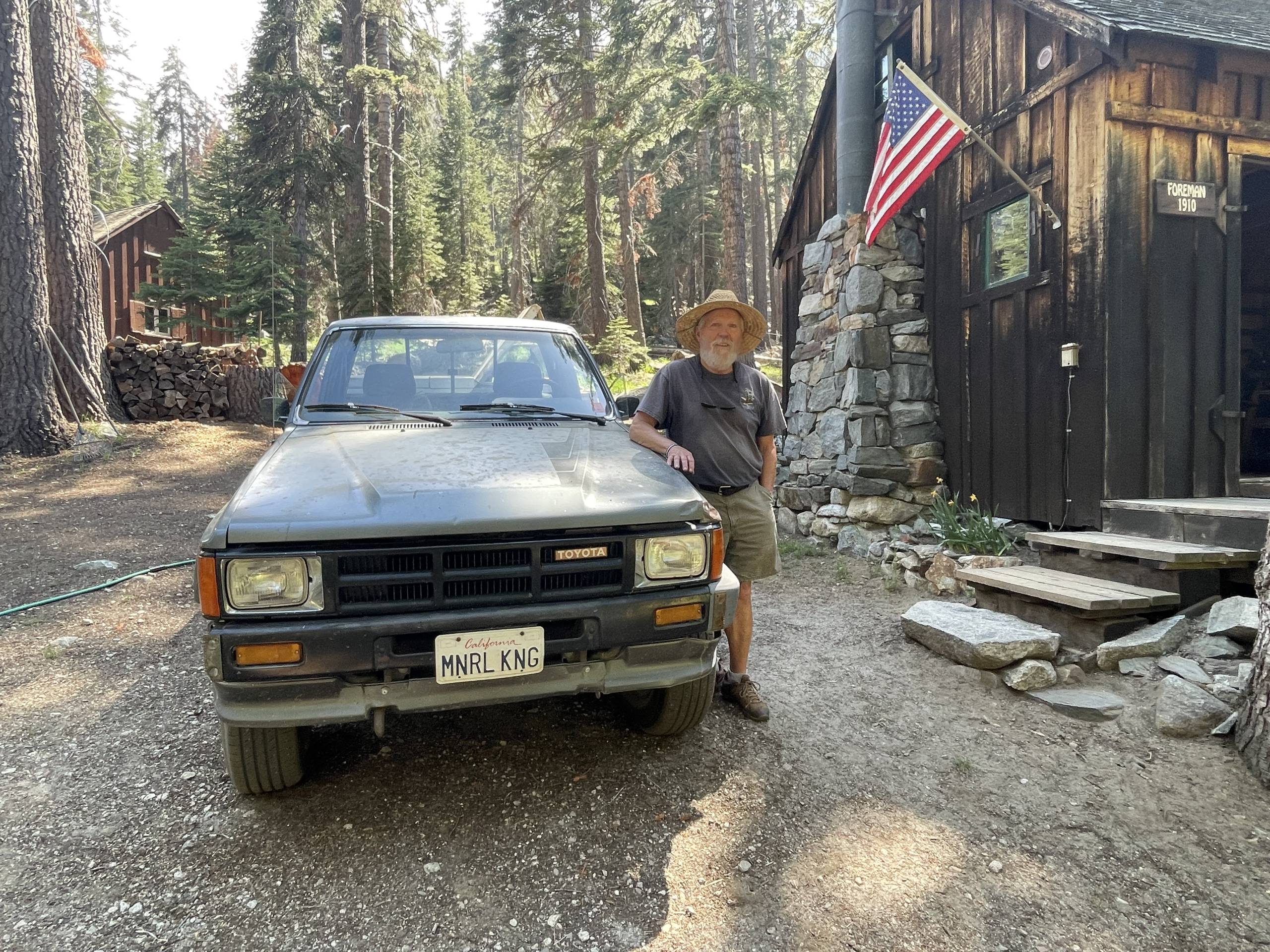 A white man wearing a hat and gray shirt rests his arm on a truck next to a cabin in the forest.