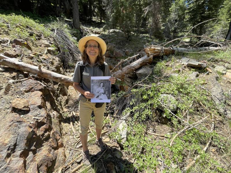 A woman wearing a hat, glasses and a green shirt holds a picture while standing in a forest.