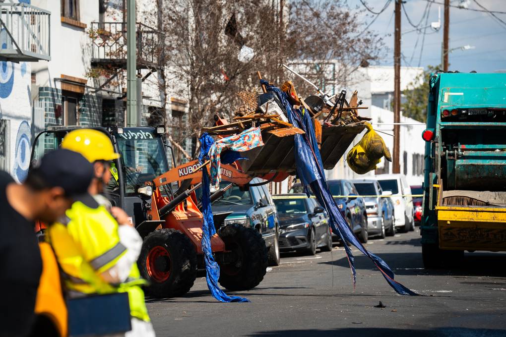 A construction vehicle lifts debris and a tent on the street with a person wearing a yellow safety helmet and bright clothing.