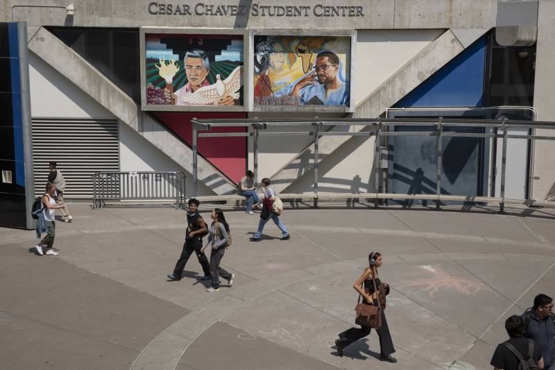 Several walk in a concrete courtyard with a sign in the background saying 'Cesar Chavez Student Center.'