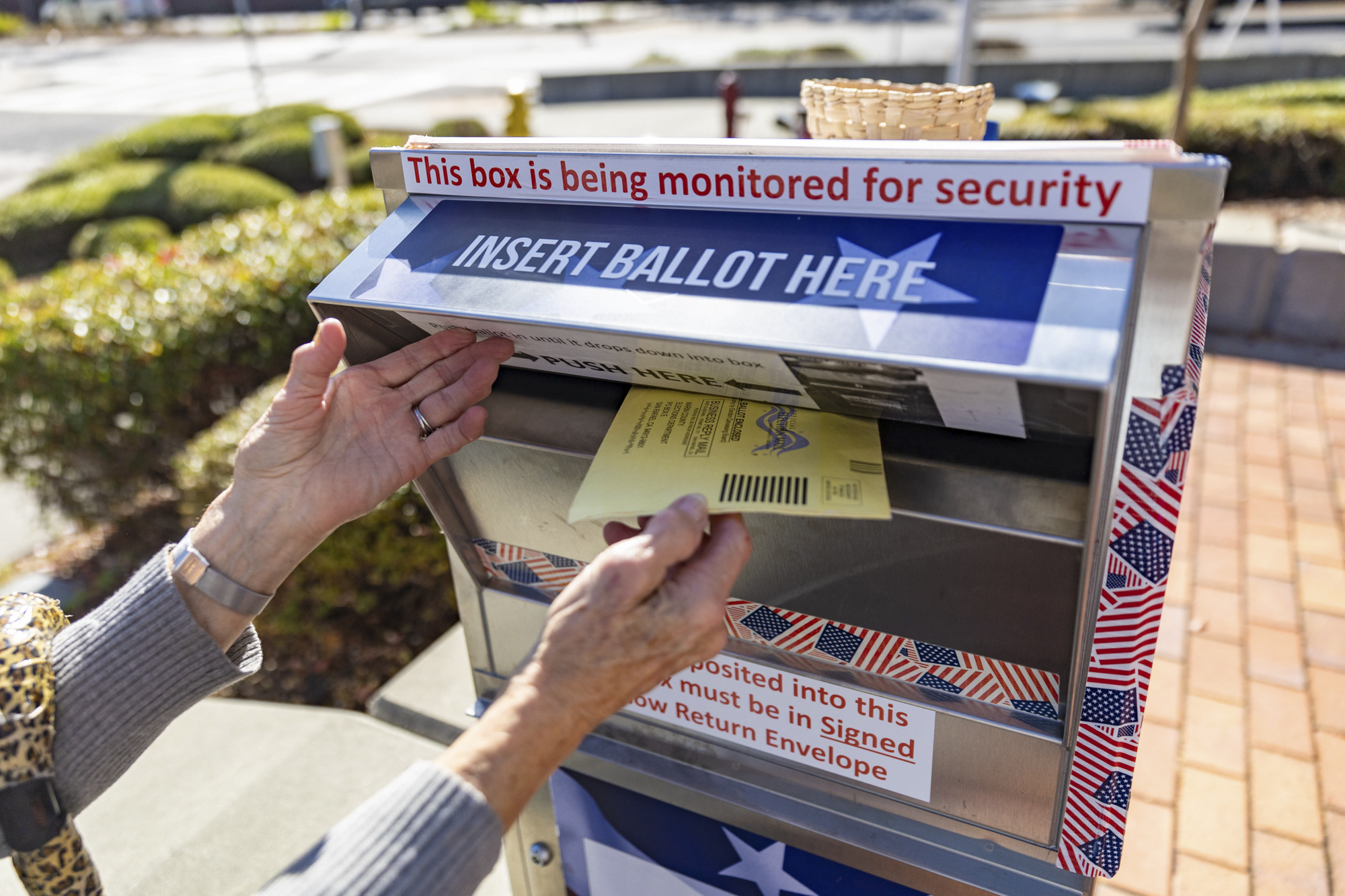 A person puts a yellow ballot envelope in a ballot drop box.
