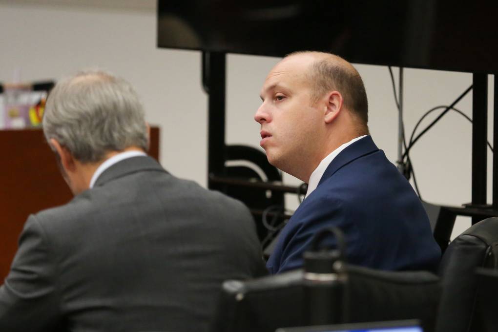 A bald, white man in a suit jacket sits in a courtroom next to another man in a suit, who is presumably is lawyer.