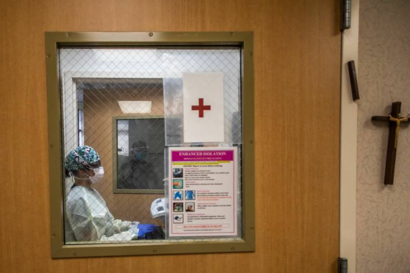 A nurse in blue scrubs seen through a small door window with a red cross posted on it.